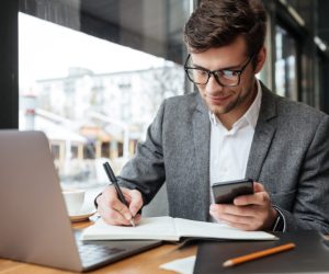 Smiling business man in eyeglasses sitting by the table in cafe with laptop computer while using smartphone and writing something