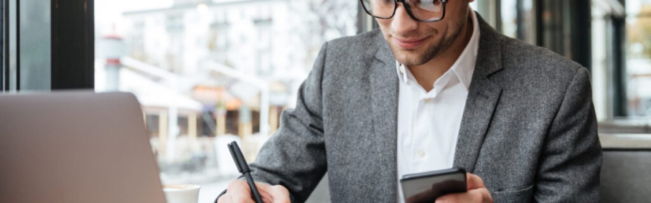 Smiling business man in eyeglasses sitting by the table in cafe with laptop computer while using smartphone and writing something